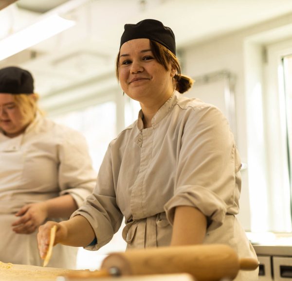 Två elever med svarta bagarmössor arbetar i bageriet, den ena ler mot kameran.